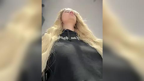 Media: Video of a woman with long blonde hair and fair skin, wearing a black shirt with a gold chain necklace, standing indoors with a blurred background.