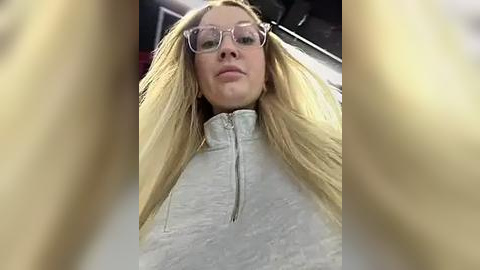 Media: Video of a blonde woman with fair skin, wearing clear glasses and a white, high-neck, textured jacket. She is indoors, with blurred, yellowish reflections framing the image.