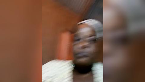 Media: A blurry video of an African woman with dark skin, wearing a light-colored headscarf and a beige shirt, standing against an orange wall. The image has a motion blur effect, making her features and surroundings indistinct.