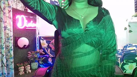Media: Video of a woman with light skin, medium build, wearing a green, shiny satin blouse, standing in a cozy room with a neon \"D\" sign, plush toys, and a bed with floral bedding.