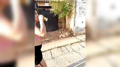 Media: A video of a woman in a pink sports bra and black shorts drinking from a can in a suburban street. She has fair skin, a small build, and is standing near a chain-link fence with a green vine and a white wall.