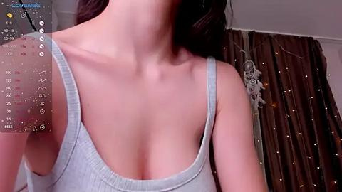 Media: Video of a woman with fair skin, wearing a light gray tank top, revealing her collarbone and cleavage. Background includes a brown curtain with string lights and a calendar overlay on the left.