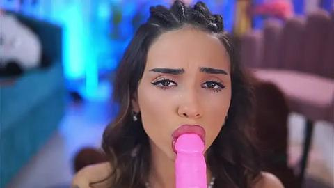 Media: Video of a young woman with light brown skin and long brown hair, wearing a pink dildo in her mouth, in a blurry, colorful, indoor setting.