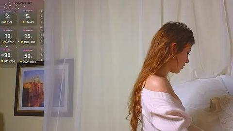 Media: Video of a woman with long, wavy brown hair in a white off-shoulder sweater, standing in a softly lit bedroom with a digital weather display, beige curtains, and a framed painting of a cityscape on the wall.