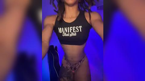 Media: Video of a muscular, light-skinned woman with dark curly hair, wearing a black sports bra with \"MANIFEST\" written in white, and camo-patterned panties. Background is a blurred, blue-lit room.