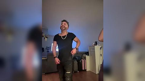 Media: Video of a middle-aged man with short brown hair, smiling, wearing a black T-shirt and jeans, standing in a dimly lit living room with wooden flooring and a beige couch.