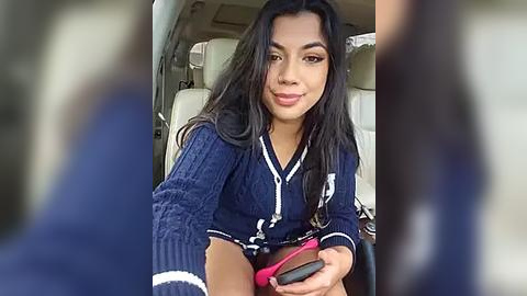 Media: Video of a young woman with long black hair and medium skin tone, wearing a navy blue cardigan and holding a pink smartphone, seated in a car with beige leather seats.