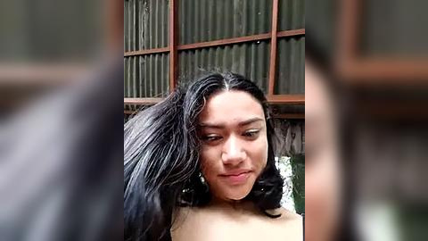 Media: Video of a young, light-skinned woman with long, wavy black hair, wearing a subtle smile, set against a rustic, wooden-framed window with corrugated metal panels in the background.