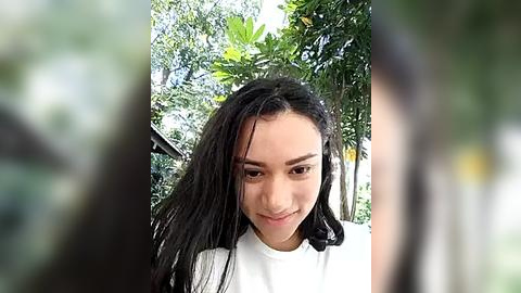 Media: Video of a young Asian woman with long black hair, wearing a white shirt, smiling outdoors against a lush green background.