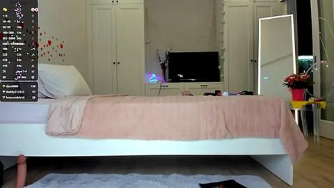 Media: Video of a minimalist bedroom with a white bed, beige blanket, and white wardrobe. A mirror and a TV stand with a black TV are visible in the background.