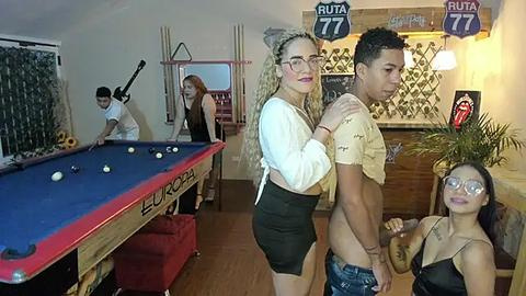 Media: Video of a lively, dimly-lit pool hall with four people: two women and two men. The women wear glasses and crop tops, while the men are shirtless. Pool table, dartboard, and sports memorabilia adorn the walls.