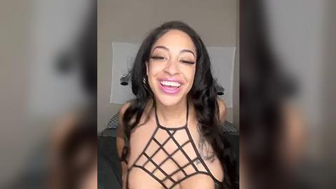 Media: A video of a joyful, light-skinned woman with long black hair, wearing a black, crisscrossed, halter top, grinning widely. The background shows a beige wall with a blurred, dark figure.