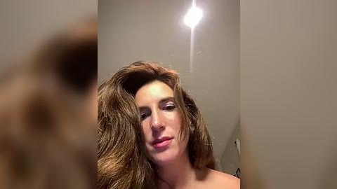 Media: Video of a woman with long, wavy brown hair, gazing into a mirror, illuminating her face with a bright overhead light. The background is a plain beige wall, and her expression is contemplative.