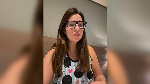 Media: Video of a Caucasian woman with long brown hair, wearing glasses and a sleeveless top with Oreo cookies design, seated indoors against a blurred background.