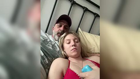 Media: A video captures a man with a beard and a woman with blonde hair lying in bed, both wearing casual clothes. The woman's red bra is visible. The background shows a black metal headboard and beige pillows.