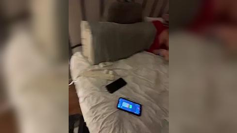 Media: Video of a messy bedroom with a gray pillow, a white pillow, a smartphone displaying a blue screen, and a black smartphone charger on a bed.