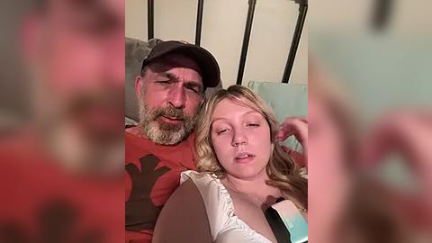 Media: Video of a middle-aged bearded man with a red shirt and cap, and a young blonde girl with a white top, lying on a bed, blurred background.