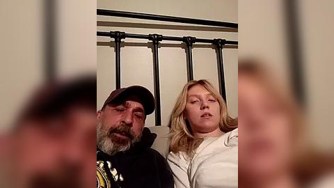 Media: A video captures a middle-aged man with a beard and a baseball cap, and a young woman with blonde hair, sitting on a bed with a black metal headboard. The image is slightly blurred.