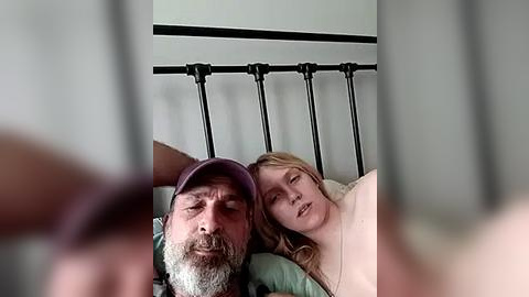 Media: Video of an older man with a grey beard and cap lying next to a blonde woman in bed, both appearing relaxed.