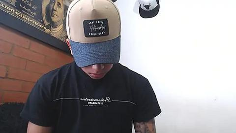 Media: Video of a young man with light skin, wearing a beige baseball cap with blue brim and a black T-shirt with white text. Background features a white wall with a mounted black and white hat and a brick wall with a framed poster.