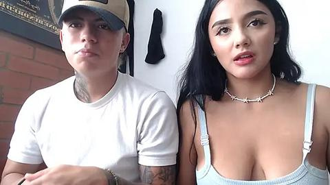 Media: Video of two people: a woman with long black hair and large breasts in a low-cut top, and a tattooed man wearing a cap and white shirt, in a brick-walled room.