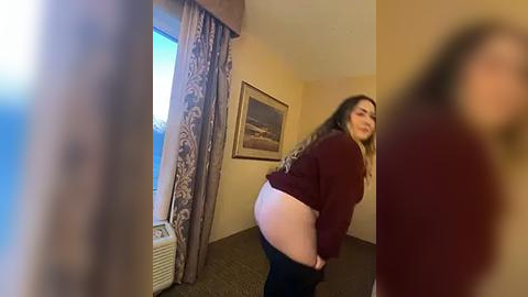 Media: Video of a plus-sized woman with long, wavy hair, wearing a maroon sweater, pulling down black pants, in a hotel room with beige walls and a framed landscape painting.