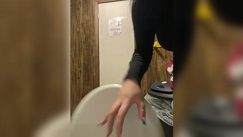 Media: Video of a person in a public restroom with a blurred face, wearing a black long-sleeve shirt, washing their hands at a white sink. The background features wooden paneling and a black garbage bin.