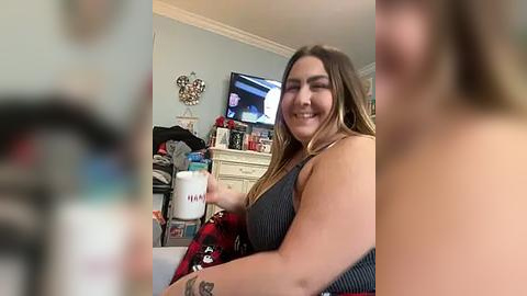 Media: Video of a smiling, light-skinned woman with long, blonde hair in a sleeveless striped dress, holding a \"Milk\" carton in a cluttered living room with a TV, bookshelf, and a wall mount.