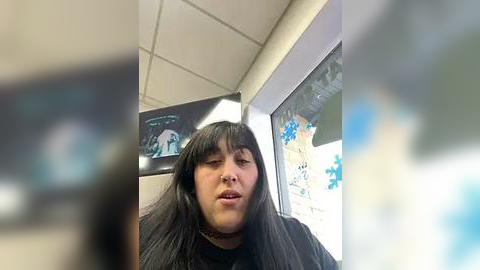 Media: Video of a woman with long black hair and bangs, wearing a black choker and a black cape, sitting in a salon with a window displaying blue and white decorations.