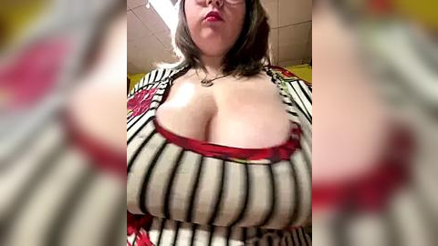 Media: Video of a plus-sized woman with fair skin and shoulder-length brown hair, wearing a low-cut, black-and-white striped top that reveals ample cleavage. Background features a yellow wall and ceiling tiles.