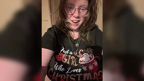 Media: Video of a smiling, fair-skinned woman with messy brown hair, wearing glasses and a Christmas-themed shirt reading \"Just a Girl Who Loves Christmas,\" indoors.