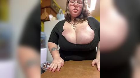 Media: Video of a plus-size woman with large breasts, wearing glasses and a black top, seated at a wooden table in a home kitchen. Background includes a fridge and cabinets.