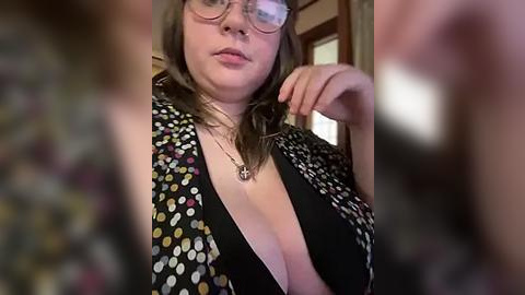 Media: A candid video of a plus-sized woman with glasses, fair skin, and shoulder-length brown hair, wearing a low-cut, patterned blouse, indoors with blurred background.