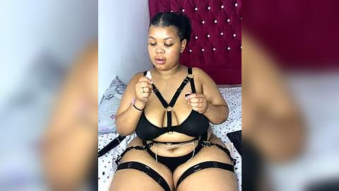 Media: Video of a curvy, dark-skinned woman with medium-length hair, wearing black strappy lingerie and garters, sitting on a bed with a tufted red headboard, in a dimly lit room.