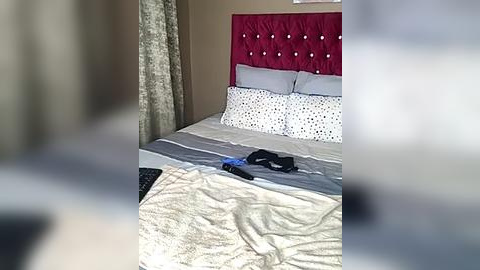 Media: A video of a neatly made bed with a tufted red headboard, white pillows, and beige bedding. A blue gaming controller and a black smartphone lie on the bed, adding a modern touch to the room.