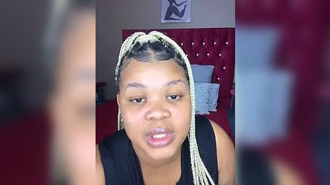 Media: Video of a Black woman with light skin, wearing braids and a black sleeveless top, sitting on a bed with a red headboard and white pillows.