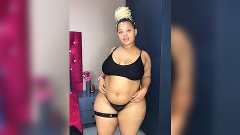 Media: A video of a plus-sized Black woman with light brown skin, wearing black lingerie, with her hair in a blonde bun. She stands in a modern bedroom with a pink wall, a blue wardrobe, and a bedside table.