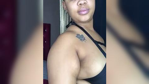 Media: Video of a light-skinned woman with braids and a black tank top, showcasing a small tattoo of a bird on her left shoulder. Background features a dark, blurry room with a red cushioned chair.