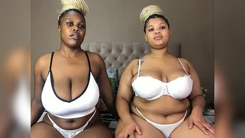 Media: Video of two African-American women with medium skin tones, wearing matching white lace lingerie, sitting on a bed with beige headboard. Both have large breasts, one with a fuller body, the other more slender.