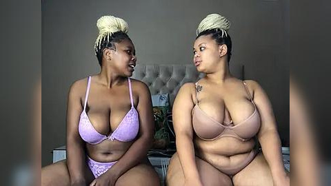 Media: Video of two African American women with dark skin, sitting on a bed, wearing matching pastel pink and beige lace lingerie. They have braids styled in updos, with one woman wearing a tattoo on her right shoulder.