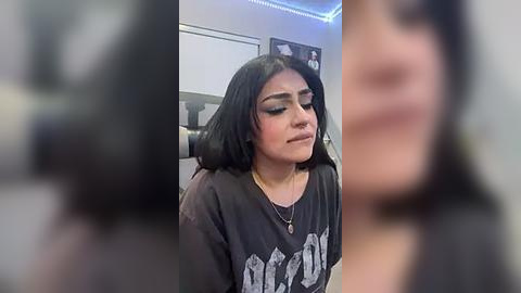 Media: Video of a young woman with long black hair, wearing heavy black eyeliner, a black graphic tee, and a necklace, sitting in a dimly lit room with blue LED lights.