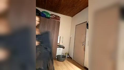 Media: Video of a messy room with a brick ceiling, a dark wooden door, a blue and green backpack on a bed, and a black trash bin beside a white cabinet.