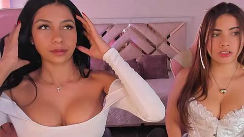 Media: Video of two young women with light brown skin, one with long black hair, the other with long brown hair. Both wear white off-shoulder tops, showcasing ample cleavage, in a modern bedroom with a plush bed and pastel decor.