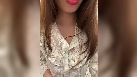 Media: A close-up video of a woman with long, straight brown hair, wearing a white lace blouse with floral patterns, and bright pink lipstick. Her face is slightly out of focus.