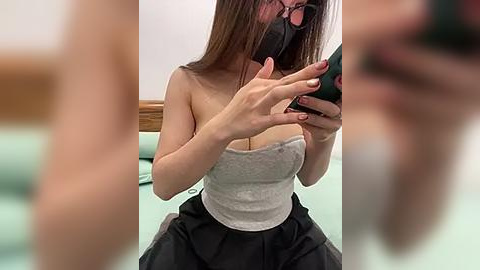 Media: Video of an East Asian woman with straight brown hair, wearing a black face mask, gray tube top, and black skirt, taking a selfie in a bathroom with a wooden bench and green tile floor.