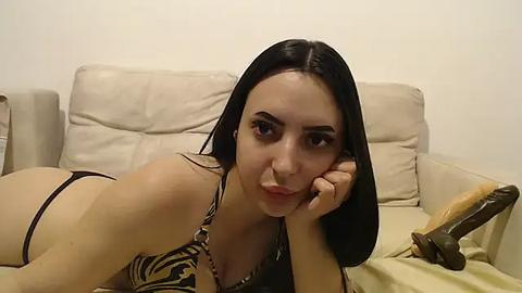 Media: A video of a young woman with long black hair, lying on a beige couch, wearing a leopard print bikini, holding a dildo.