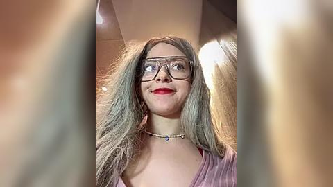 Media: Video of a woman with long, wavy gray hair, wearing red lipstick, glasses, and a pink striped top, standing in a dimly lit room with blurred figures in the foreground.