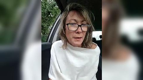 Media: Video of a middle-aged woman with glasses, wearing a white turtleneck, sitting in a car with green foliage in the background. She appears to be speaking, with blurred out faces in the foreground.