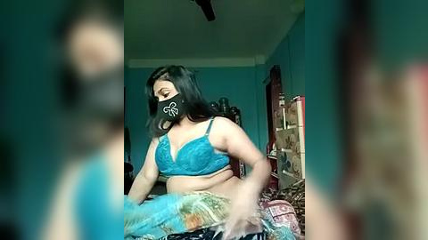 Media: Video of a plus-sized woman in a teal bra and black face mask, sitting on a bed, with blurred figures in the foreground.