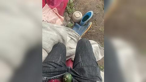 Media: Video of a person's legs in dark denim jeans, sitting on a red blanket outdoors. A blue flip-flop is nearby, and a white cloth is folded beside them. Background includes a dirt path and scattered greenery.